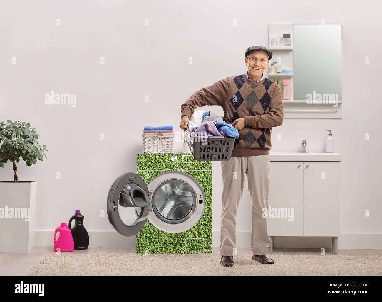 Uomo anziano sorridente che tiene in mano un cesto per lavanderia pieno di vestiti all'interno di un bagno con una lavatrice ad alta efficienza energetica Foto Stock