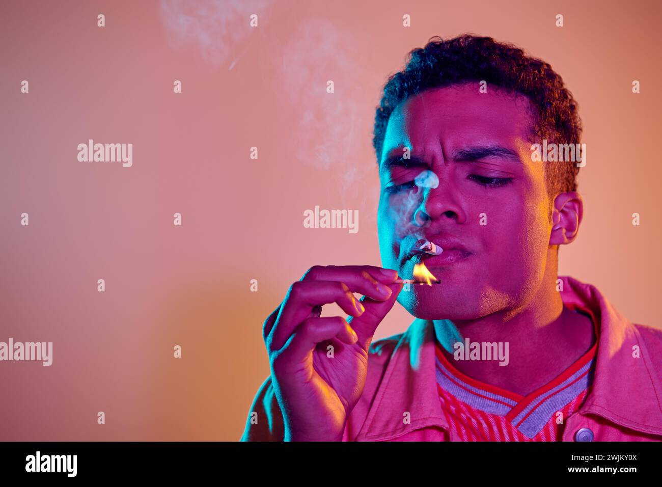 giovane uomo nero che accende la sigaretta con fiammifero sotto luci al neon blu su sfondo rosa Foto Stock