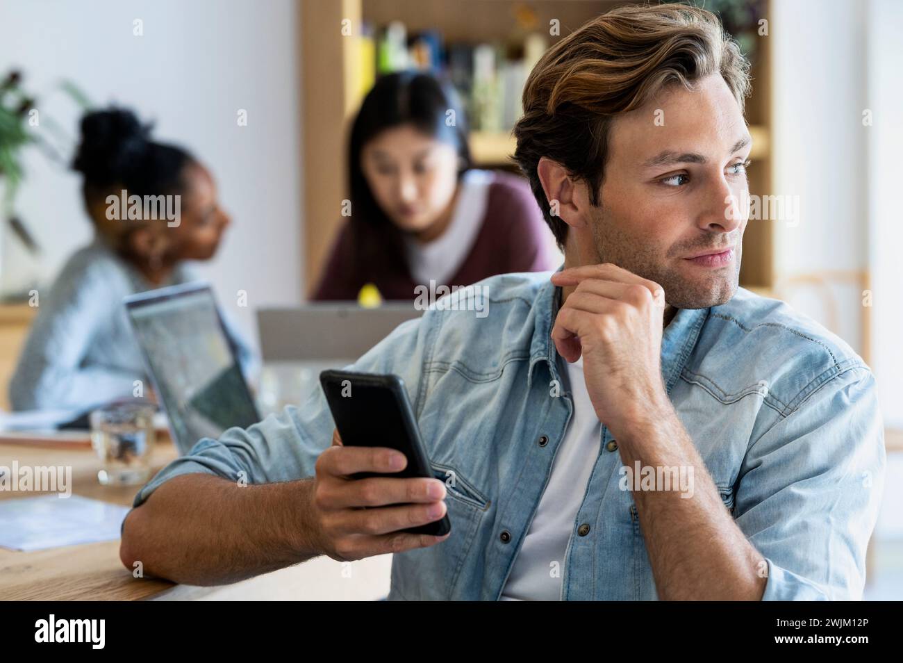 Giovane imprenditore adulto che tiene in mano uno smartphone con colleghi in background Foto Stock
