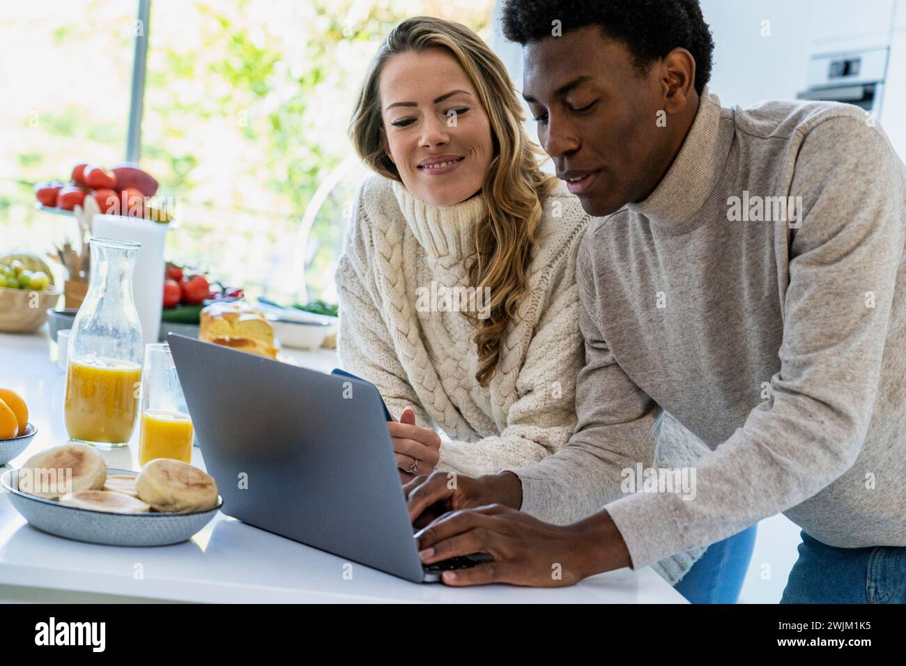 Uomo adulto che scrive sul computer portatile mentre si appoggia al bancone della cucina con la fidanzata Foto Stock