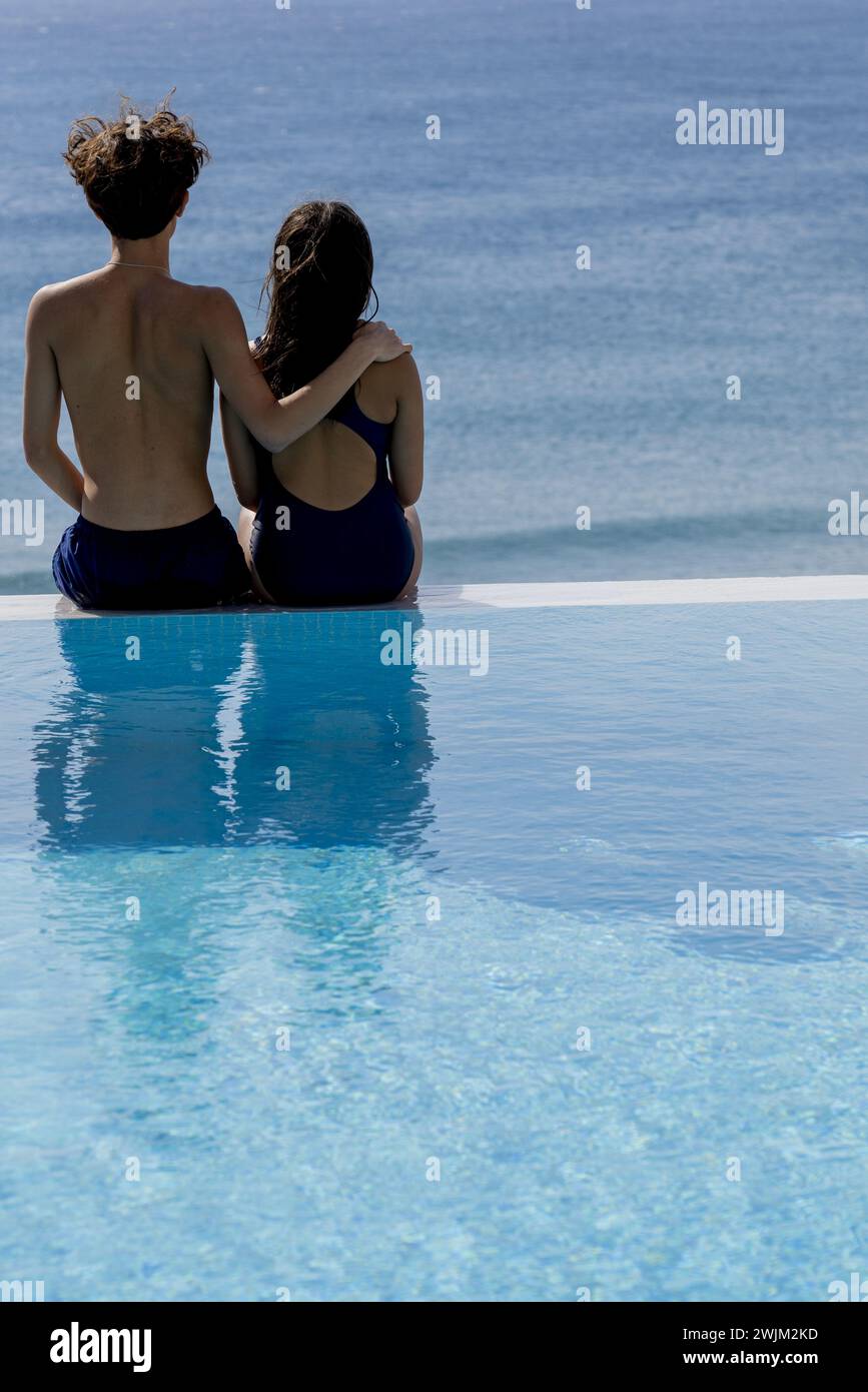 Vista posteriore di giovani coppie che indossano un costume da bagno sedute sul bordo della piscina e guardano il mare Foto Stock