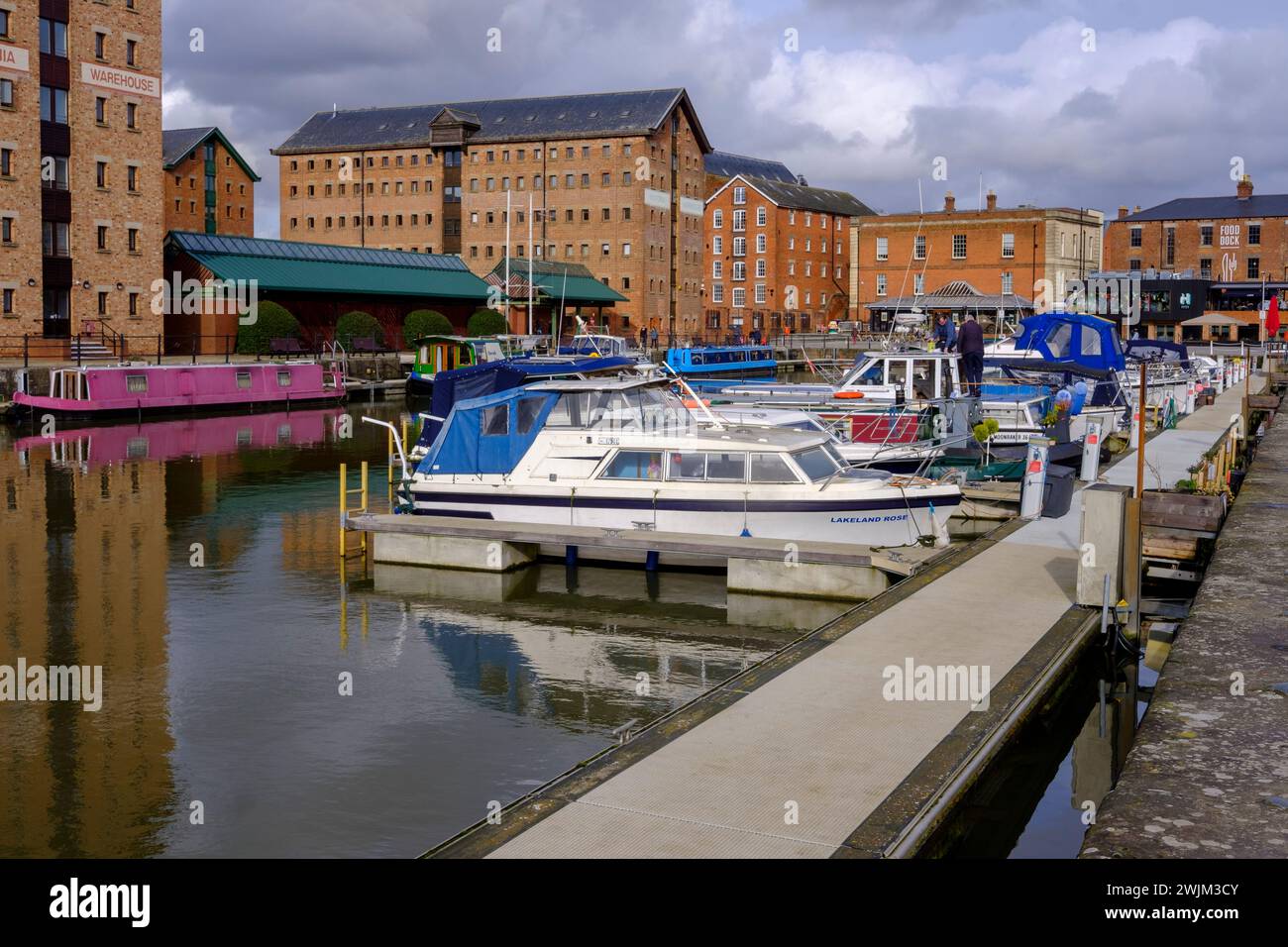 Serena vista del Gloucester Docks, con barche ormeggiate lungo il molo, con storici edifici in mattoni sullo sfondo sotto un cielo parzialmente nuvoloso Foto Stock