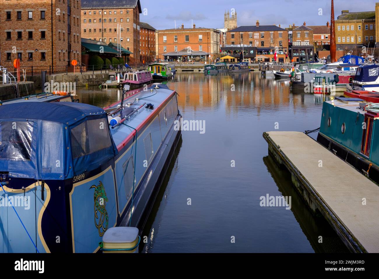 Serena vista del Gloucester Docks, con barche ormeggiate lungo il molo, con storici edifici in mattoni sullo sfondo sotto un cielo parzialmente nuvoloso Foto Stock