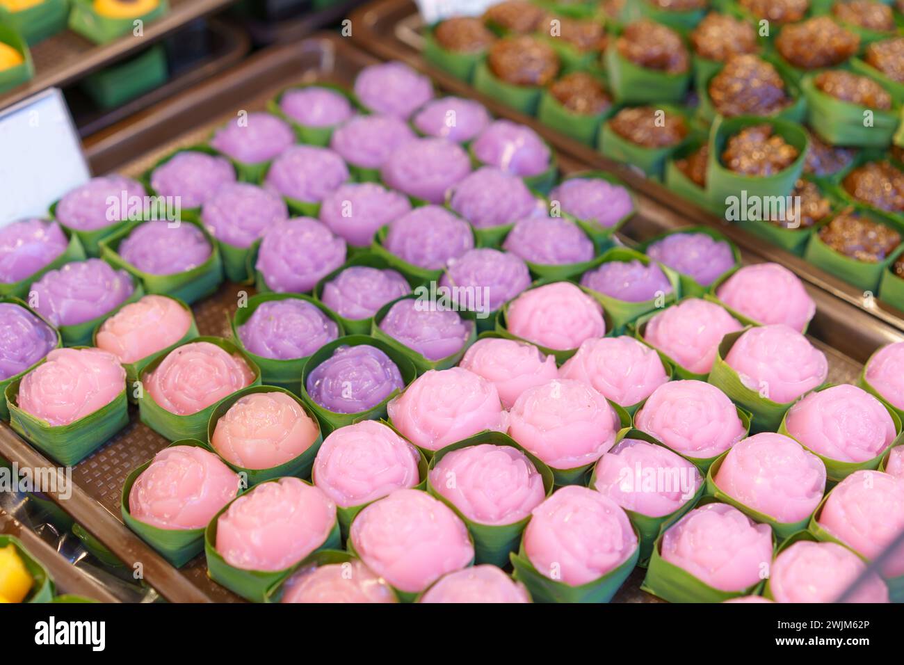 Dolci a strati cotti al vapore realizzati in forme floreali, presentati in tazze di foglie di banana, con una varietà di colori rosa e viola Foto Stock