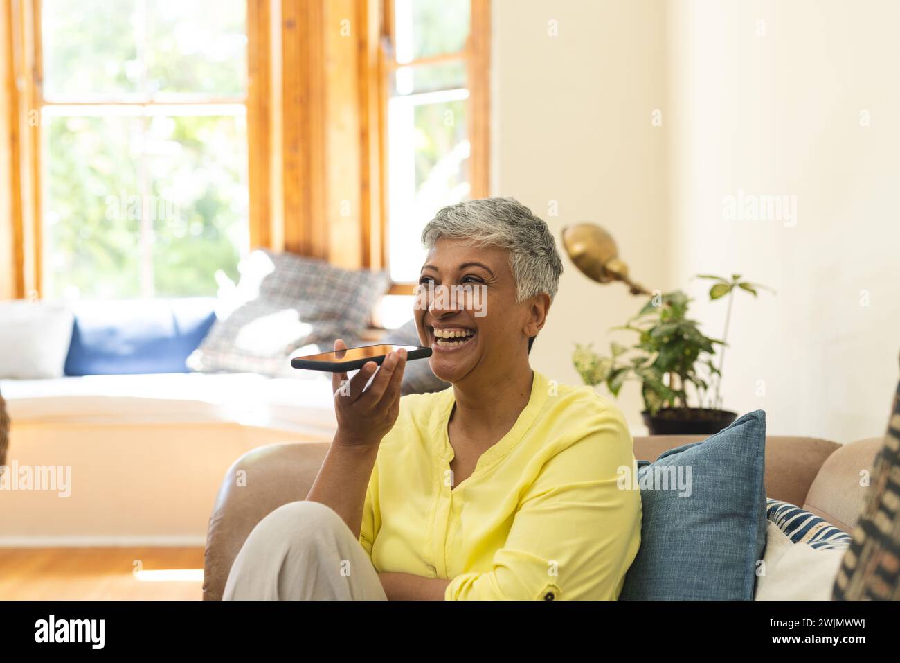 Donna matura birazziale allegra che utilizza i comandi vocali sullo smartphone a casa Foto Stock