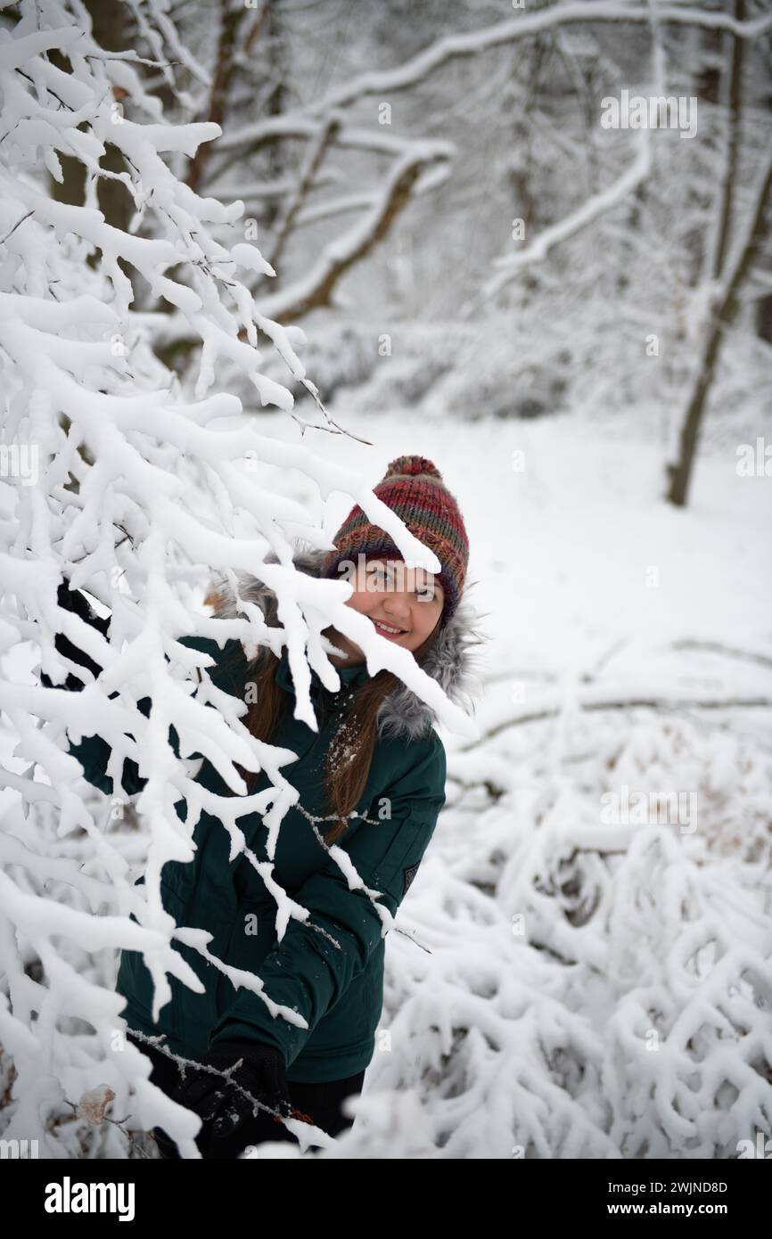 Una donna sorride mentre sbircia da dietro rami pesanti di neve in un ambiente boschivo invernale. Indossa una giacca color acqua e una h multicolore Foto Stock