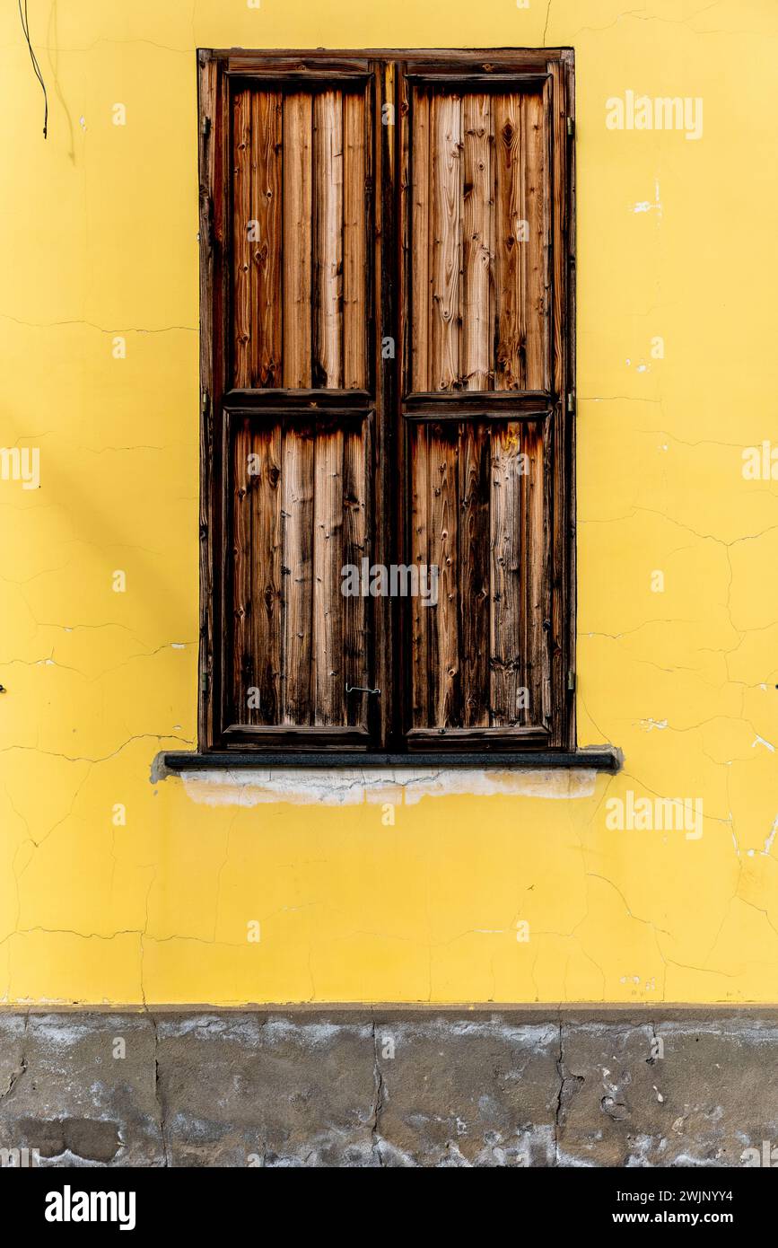 Parete della casa dipinta di giallo con superficie incrinata, due finestre. Finestra con persiane marroni su una parete gialla. Foto Stock