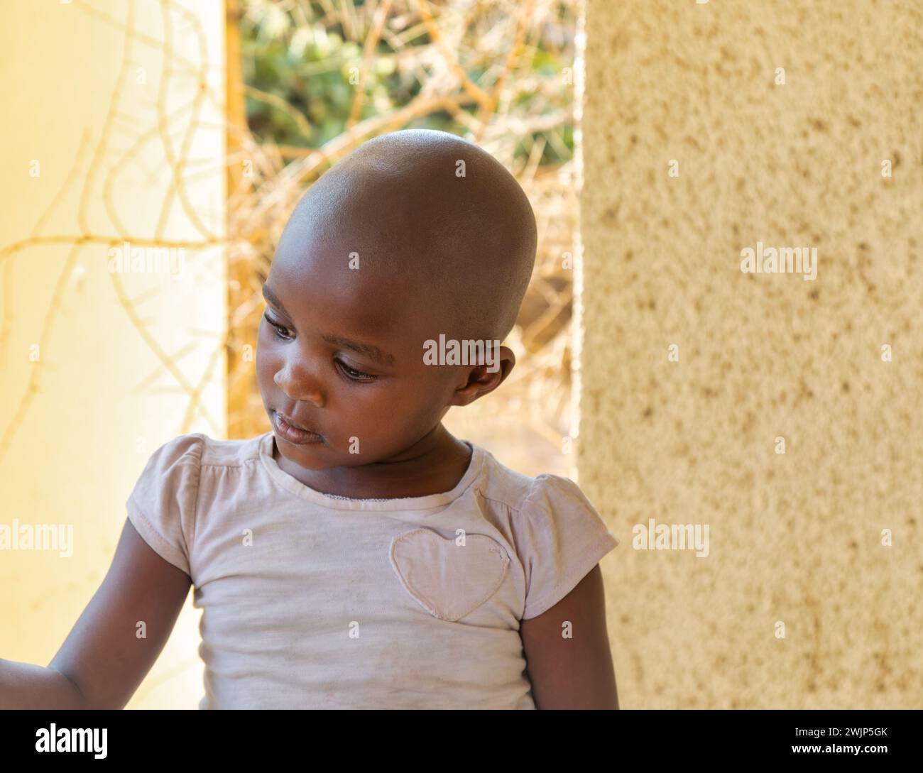 Ragazza africana del villaggio con la testa rasata, indossa una camicia rosa nel cortile Foto Stock