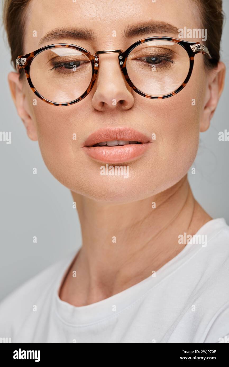 bella donna bionda con capelli raccolti e occhiali che posano sullo sfondo grigio e guardano lontano Foto Stock