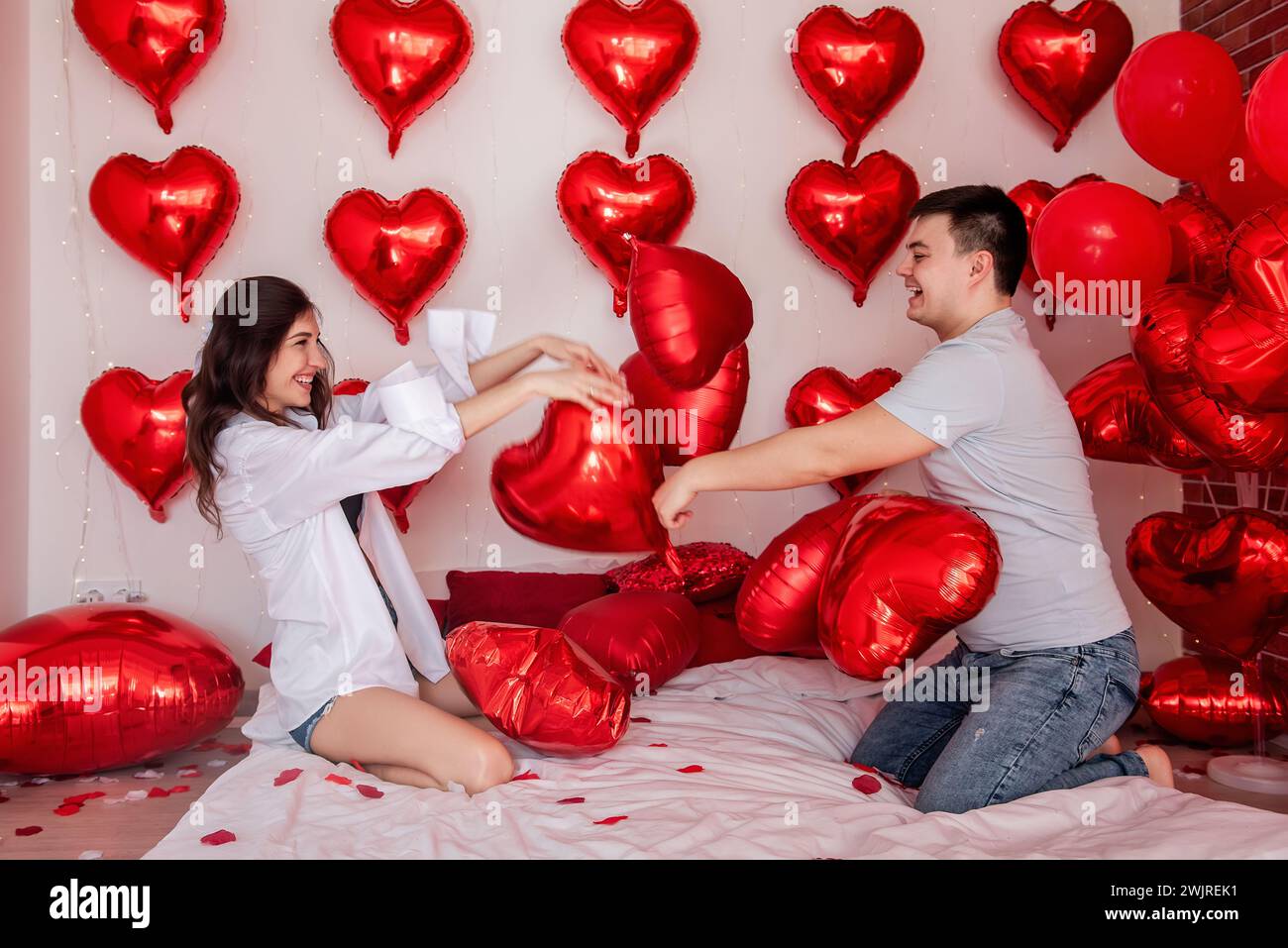 Momenti di divertimento tra le coppie che si impegnano in palloncini a cuscino che combattono sul letto, con palloncini rossi a forma di cuore sullo sfondo. La donna si sta divertendo con gioia Foto Stock