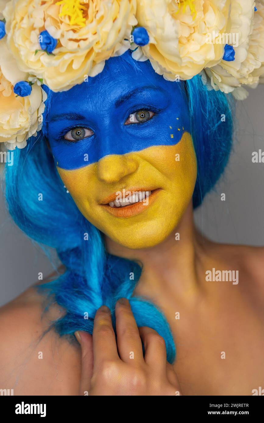 Ritratto ravvicinato creativo di una giovane donna con il viso giallo e blu, una corona di peonie sulla testa su uno sfondo grigio isolato. Concetto per Foto Stock