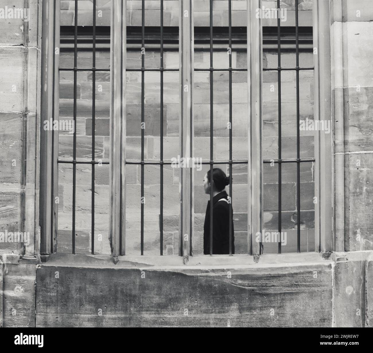 Strasburgo Francia 12.22.24 donne asiatiche stanno, guardando lontano, dietro una finestra barrata. Edificio in pietra. Capelli corti legati. Lungo cappotto nero. Posa solenne. IR Foto Stock