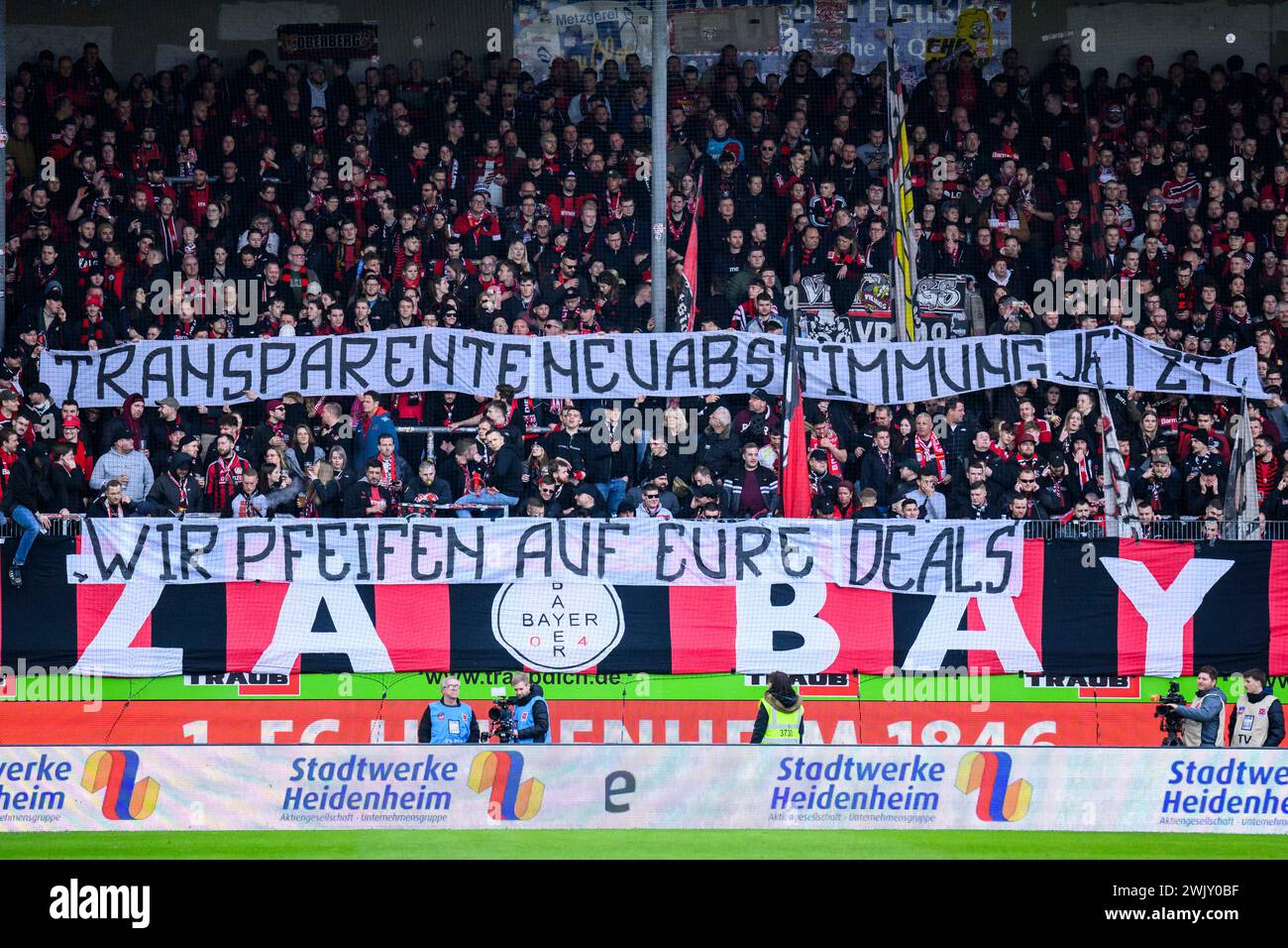 17 febbraio 2024, Baden-Württemberg, Heidenheim: Calcio: Bundesliga, 1. FC Heidenheim - Bayer Leverkusen, Matchday 22, Voith-Arena. I fan di Leverkusen tengono poster con lo slogan "Transparent Vote Now! Non ci importa niente dei vostri accordi per protestare contro i piani della Lega calcistica tedesca (DFL) di attirare investitori. Foto: Tom Weller/dpa - NOTA IMPORTANTE: In conformità con le normative della DFL German Football League e della DFB German Football Association, è vietato utilizzare o far utilizzare fotografie scattate nello stadio e/o della partita sotto forma di imag sequenziale Foto Stock