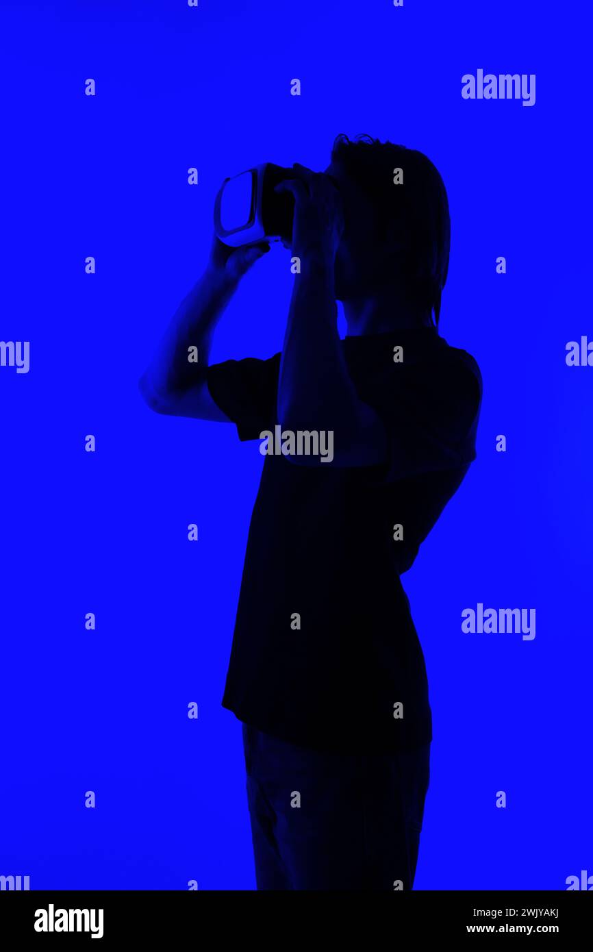 Silhouette in un giovane irriconoscibile che indossa occhiali di protezione per la realtà virtuale su uno sfondo blu luminoso, un concetto futuristico di metaversa Foto Stock