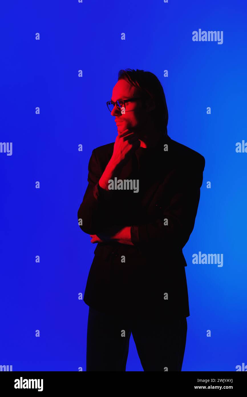 Elegante uomo d'affari con occhiali da vista in una giacca in piedi in una stanza buia con luce rossa al neon illuminata sul viso su sfondo blu Foto Stock