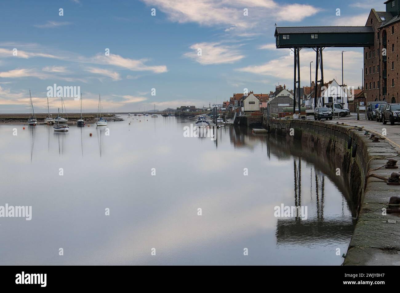 Wells-NEXT-the-Sea, Norfolk, Inghilterra, Regno Unito - barche a vela ormeggiate nelle acque calme del porto durante l'alta marea. Edificio di granaio e cielo riflesso nell'acqua Foto Stock