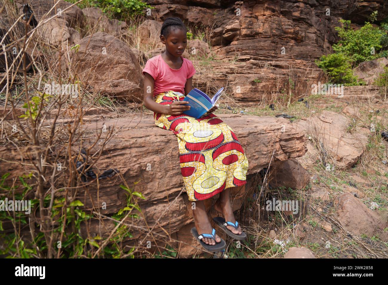 Giovane studentessa elementare nera seduta su una roccia leggendo nel suo libro di testo - concetto di educazione femminile e empowerment in Africa Foto Stock