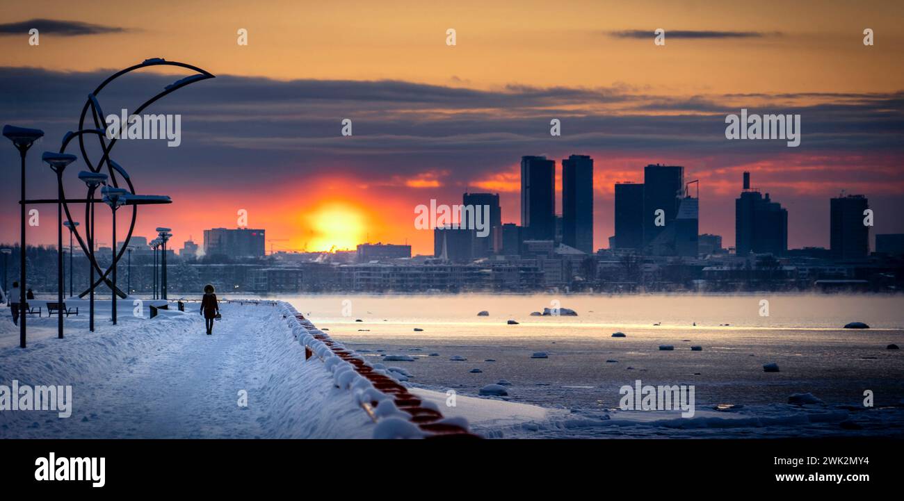 Una persona che cammina sulla passeggiata di Pirita guardando lo skyline di Tallinn in un tramonto invernale. Mare quasi ghiacciato sul lato destro, neve sul marciapiede. Foto Stock