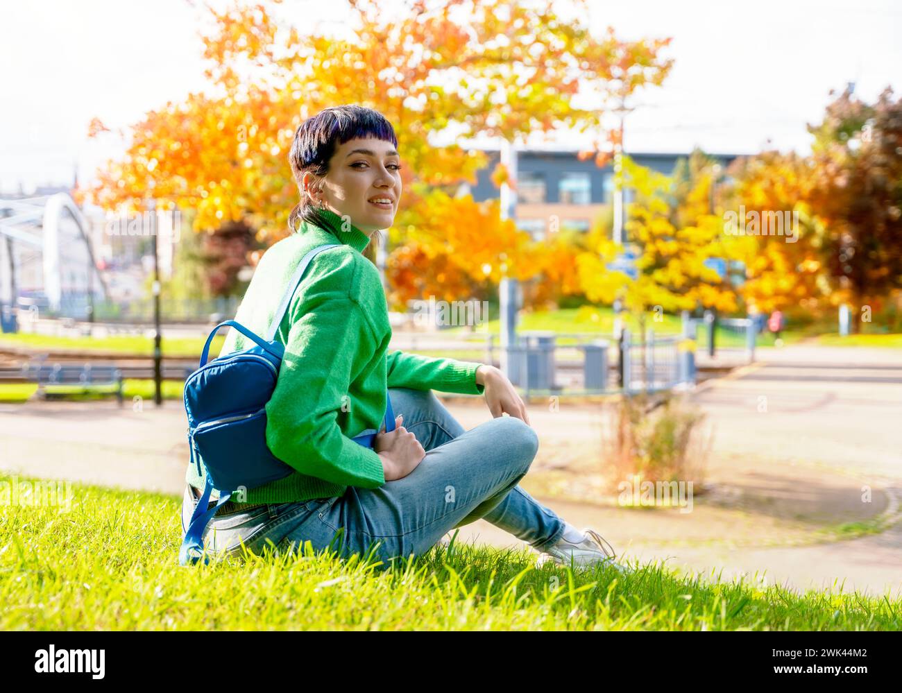 Giovane donna sorridente in maglione verde con uno zaino blu seduta nel parco sull'erba in una giornata di sole autunnale donna senza pensieri che si sente destressante Foto Stock