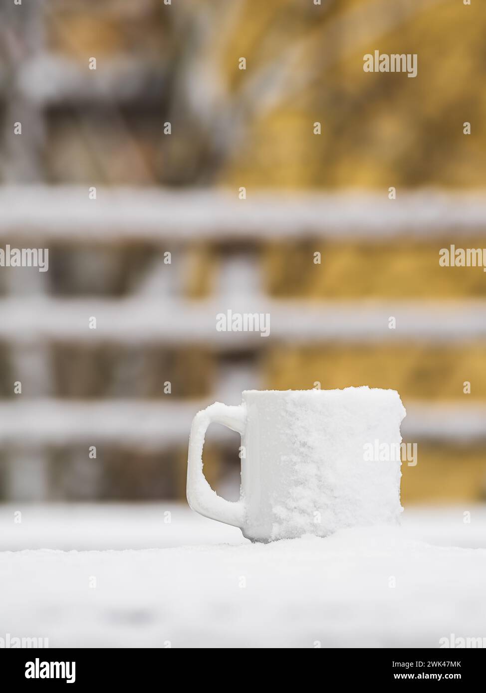 In questa tranquilla scena, una tazza bianca ricoperta di neve fresca si trova in cima a una ringhiera di legno, che offre un netto contrasto con le foglie autunnali gialle che un Foto Stock