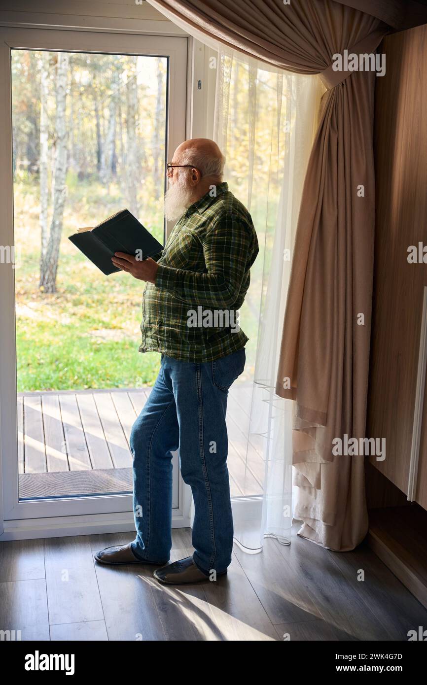 Il vecchio sta con un libro alla finestra francese Foto Stock