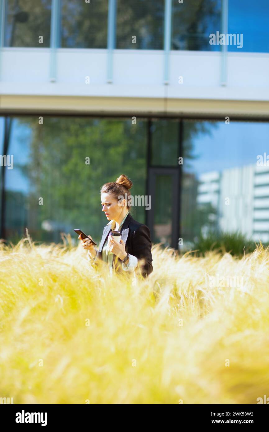 donna d'affari moderna di 40 anni, vicino al business center con giacca nera con smartphone e tazza di caffè. Foto Stock
