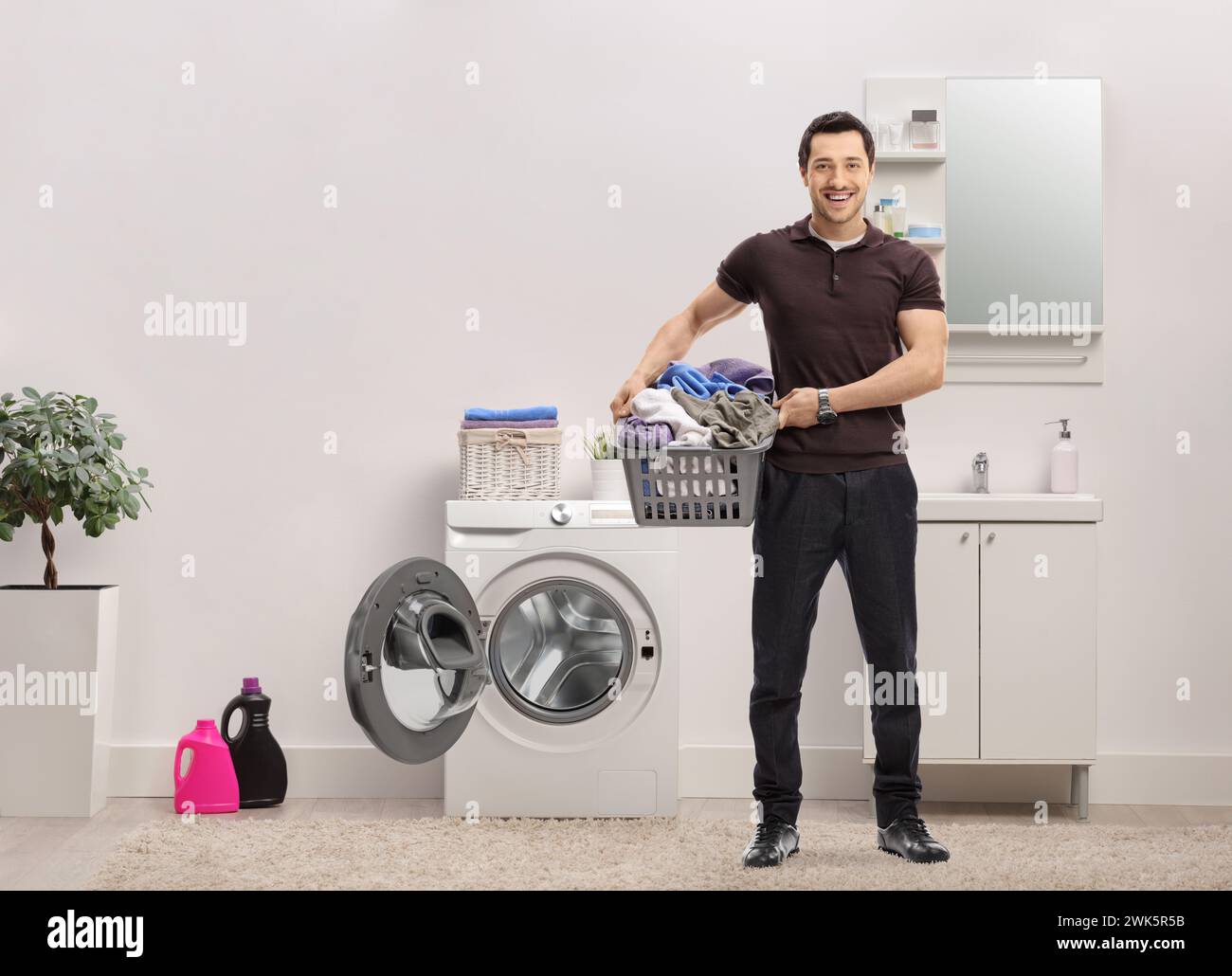 Giovane uomo con un cesto per bucato pieno di vestiti in piedi in un bagno accanto a una lavatrice Foto Stock