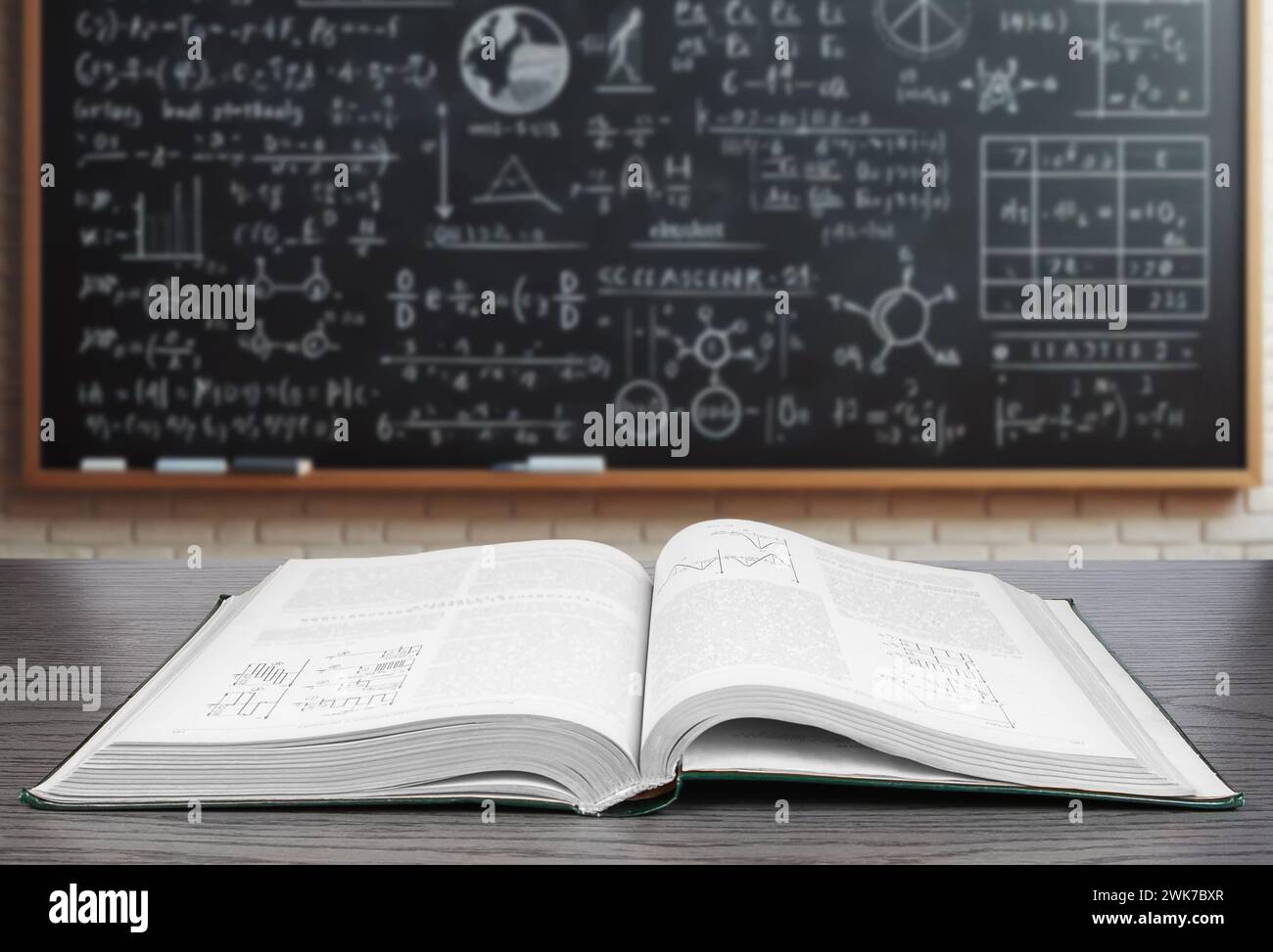Un libro di testo aperto giace su una scrivania di legno in primo piano con uno sfondo sfocato caratterizzato da una lavagna coperta da varie equazioni matematiche Foto Stock