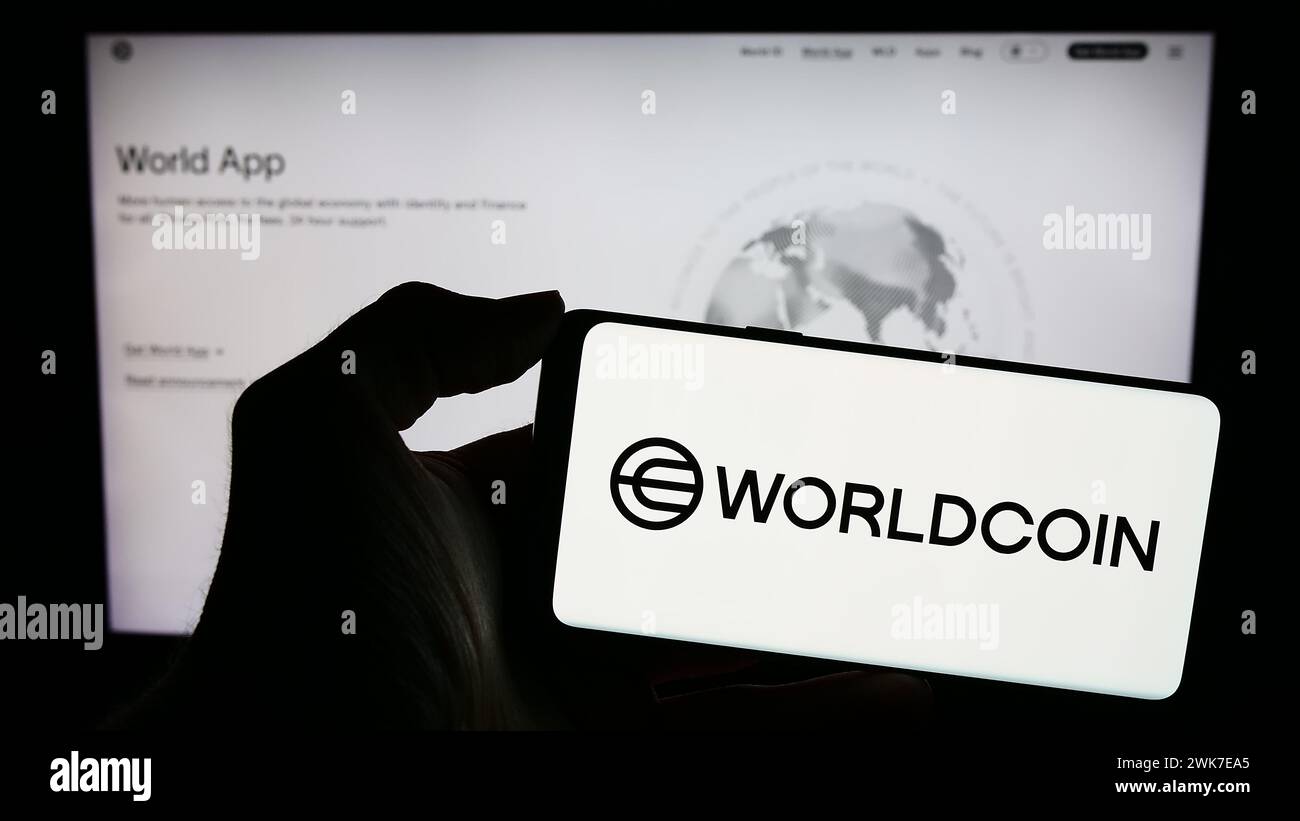 Persona che possiede un telefono cellulare con il logo del progetto di criptovaluta Worldcoin (Tools for Humanity) davanti alla pagina web. Mettere a fuoco il display del telefono. Foto Stock