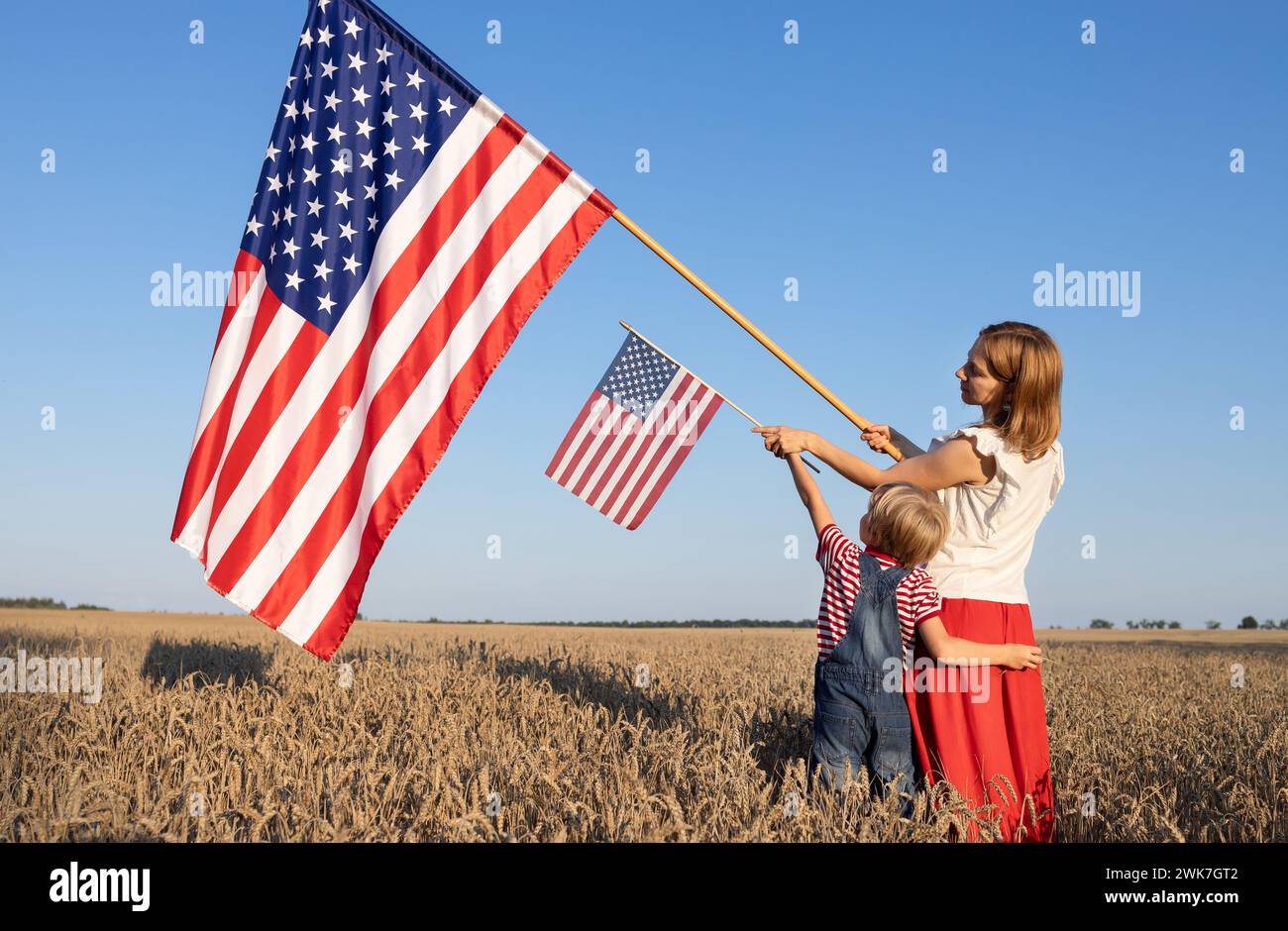 Grande e piccola bandiera americana nelle mani di una donna e di un bambino in piedi in un campo di grano illuminato dalla luce del sole. Giorno dell'indipendenza della United St Foto Stock