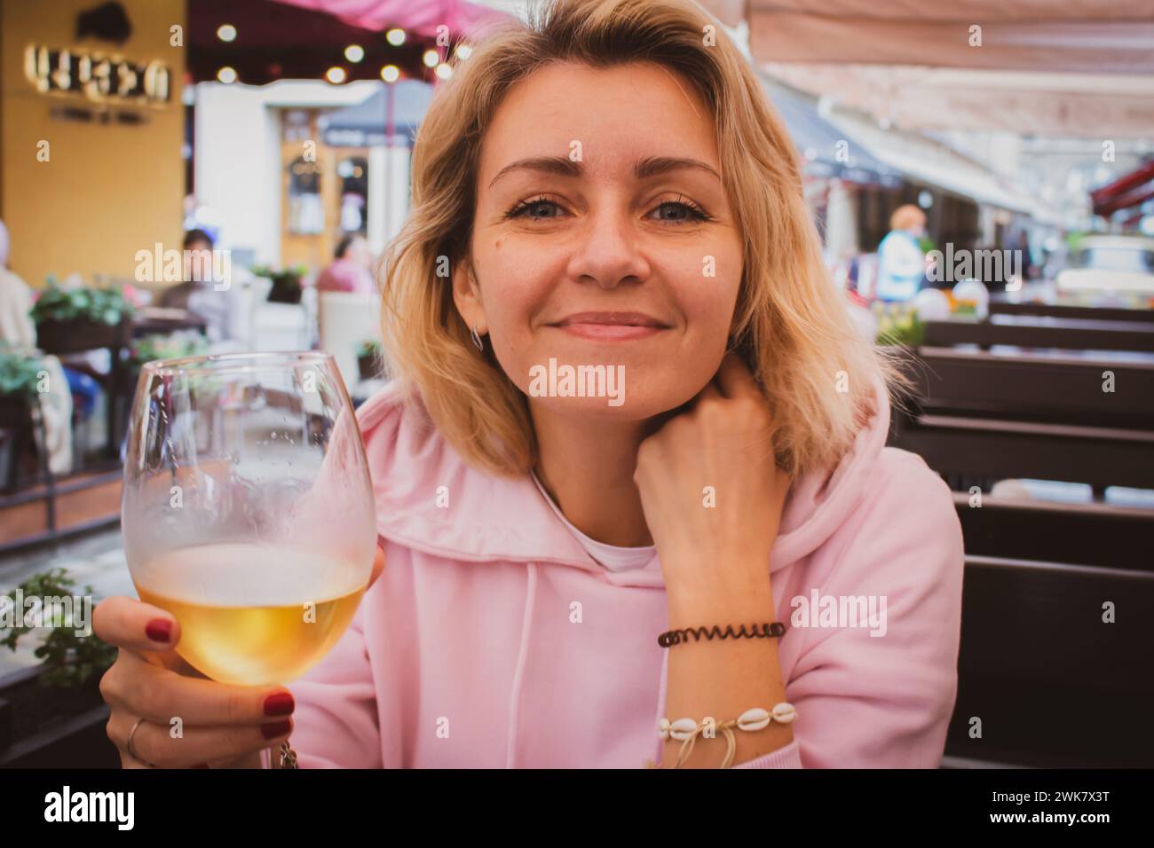 Donna sorridente con un bicchiere di vino bianco che guarda la macchina fotografica. Ragazza felice con vino nel caffè all'aperto. Stili di vita urbani. Donna al ristorante marciapiede. Foto Stock