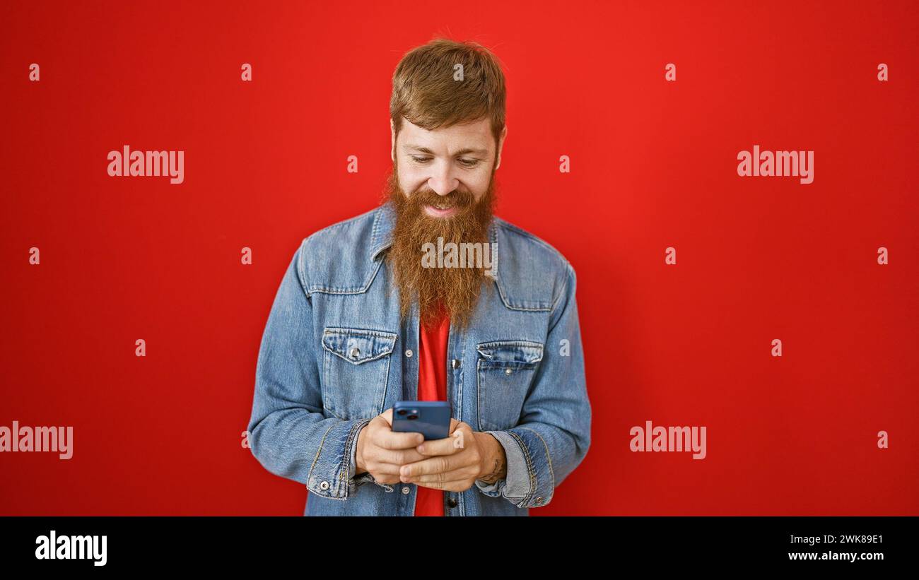 Bellissimo giovane uomo rosso, traboccante di felicità, digitando con sicurezza i messaggi sul suo smartphone su sfondo rosso isolato. la sua risata e.. Foto Stock