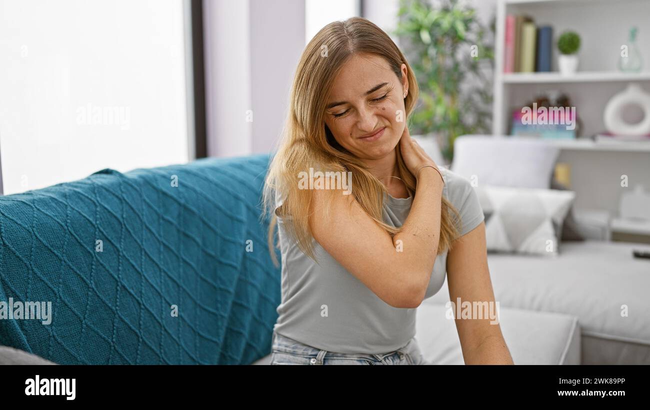 Stanca giovane donna bionda seduta sul divano soffre di intenso dolore cervicale in casa Foto Stock