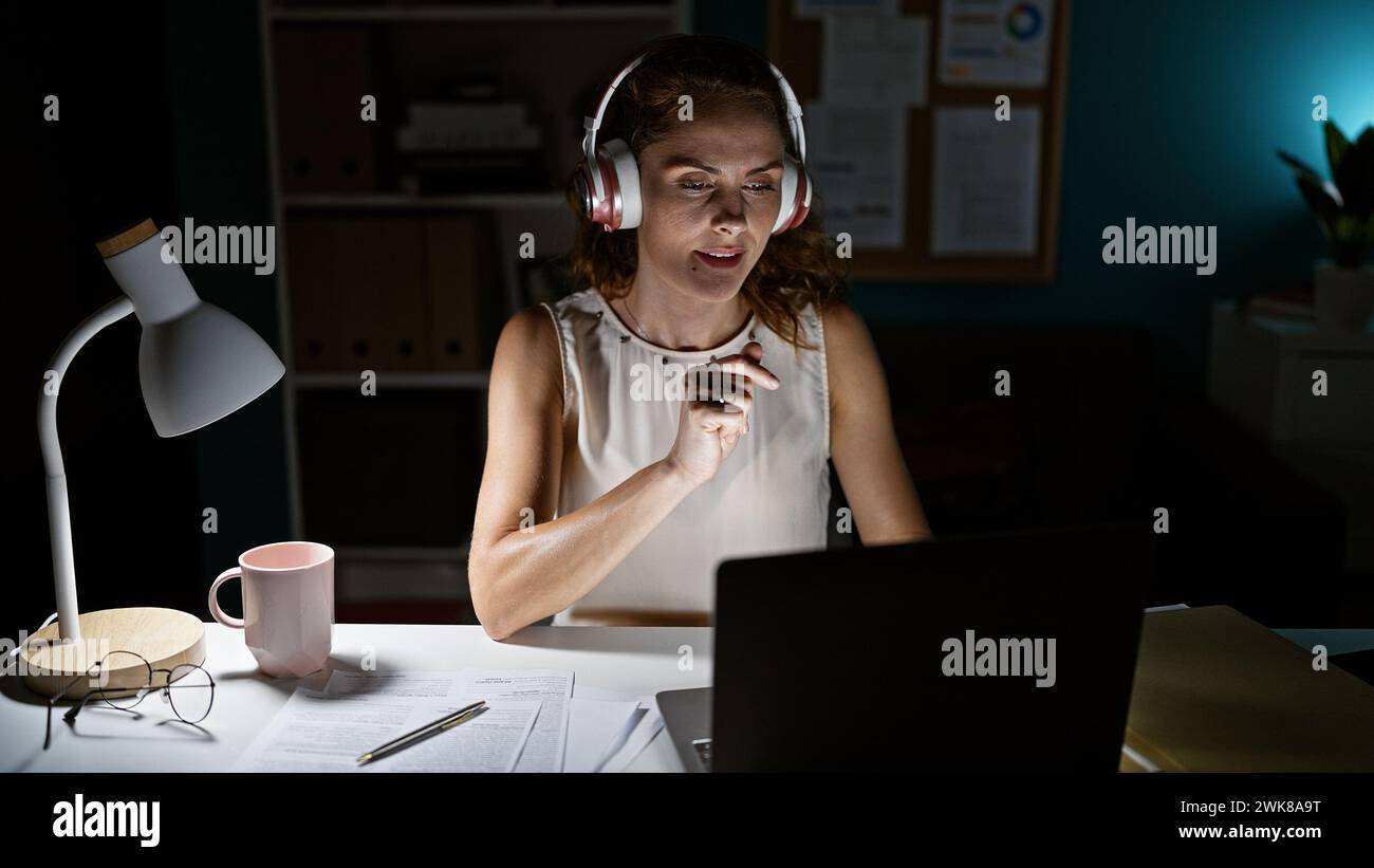 Giovane donna concentrata con le cuffie che lavora fino a tardi in un ufficio domestico illuminato da una lampada da scrivania. Foto Stock