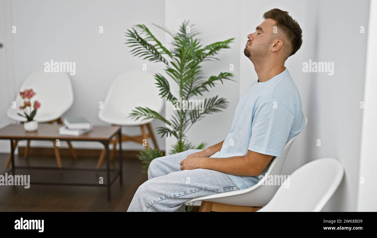 Un giovane ispanico si rilassa in una sala d'attesa dallo stile moderno con sedie bianche e piante. Foto Stock