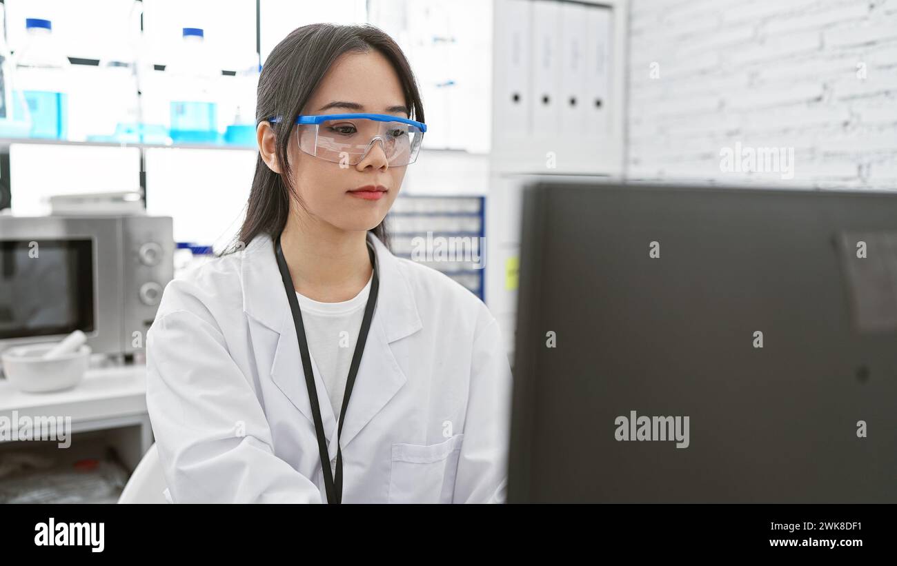 Una scienziata asiatica concentrata in un laboratorio, che indossa un camice da laboratorio bianco, occhiali di sicurezza, che analizza i dati su un computer. Foto Stock