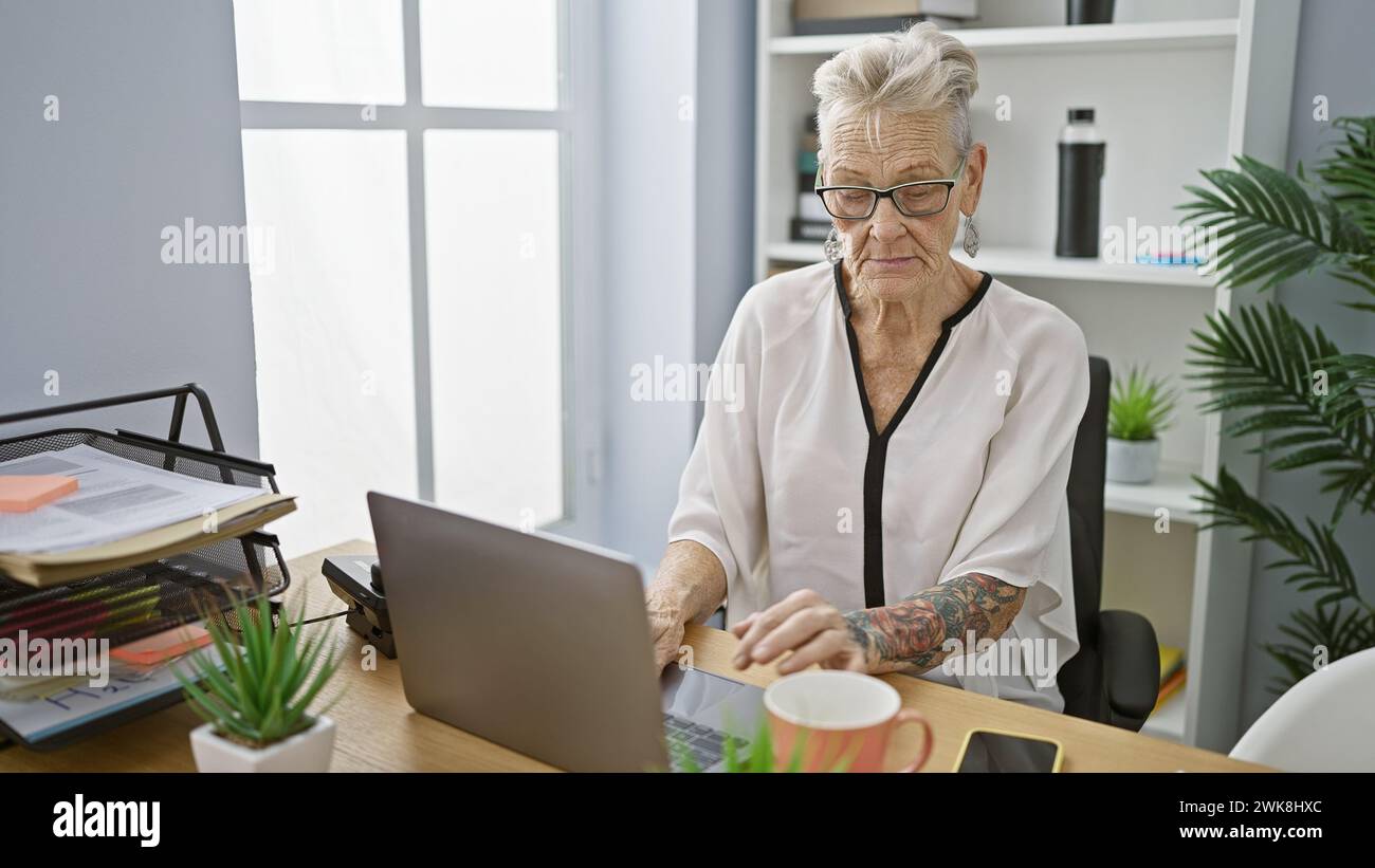 Donna anziana dai capelli grigi che lavora con zelo su un notebook mostrando il suo acume professionale in ufficio, incarnando il successo femminile Foto Stock