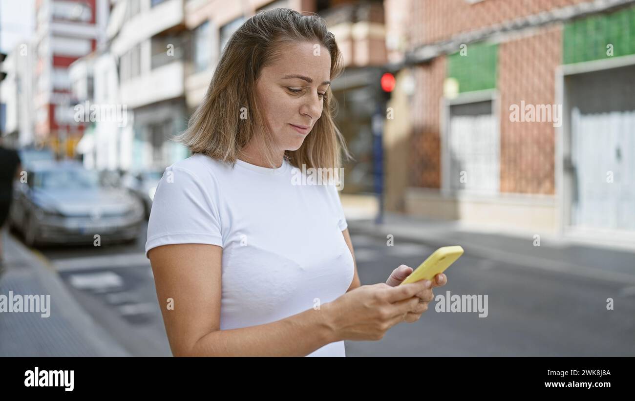 Elegante, bella e giovane donna bionda, si è concentrata seriamente sul inviare messaggi online sul suo smartphone, in piedi sulla soleggiata strada della città all'aperto, Livin Foto Stock