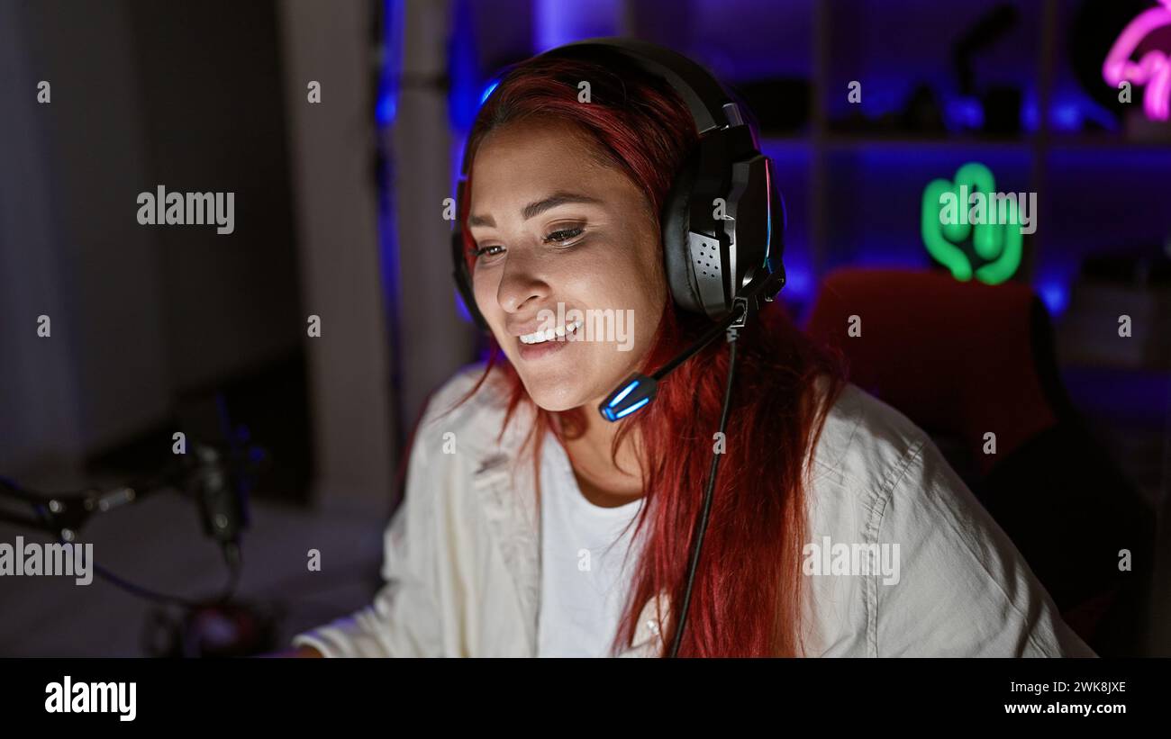 Streaming live accattivante, giovane donna che filtra rossa brillante, sorridente, che abbraccia i giochi high-tech in una stanza accogliente, con cuffie accese Foto Stock