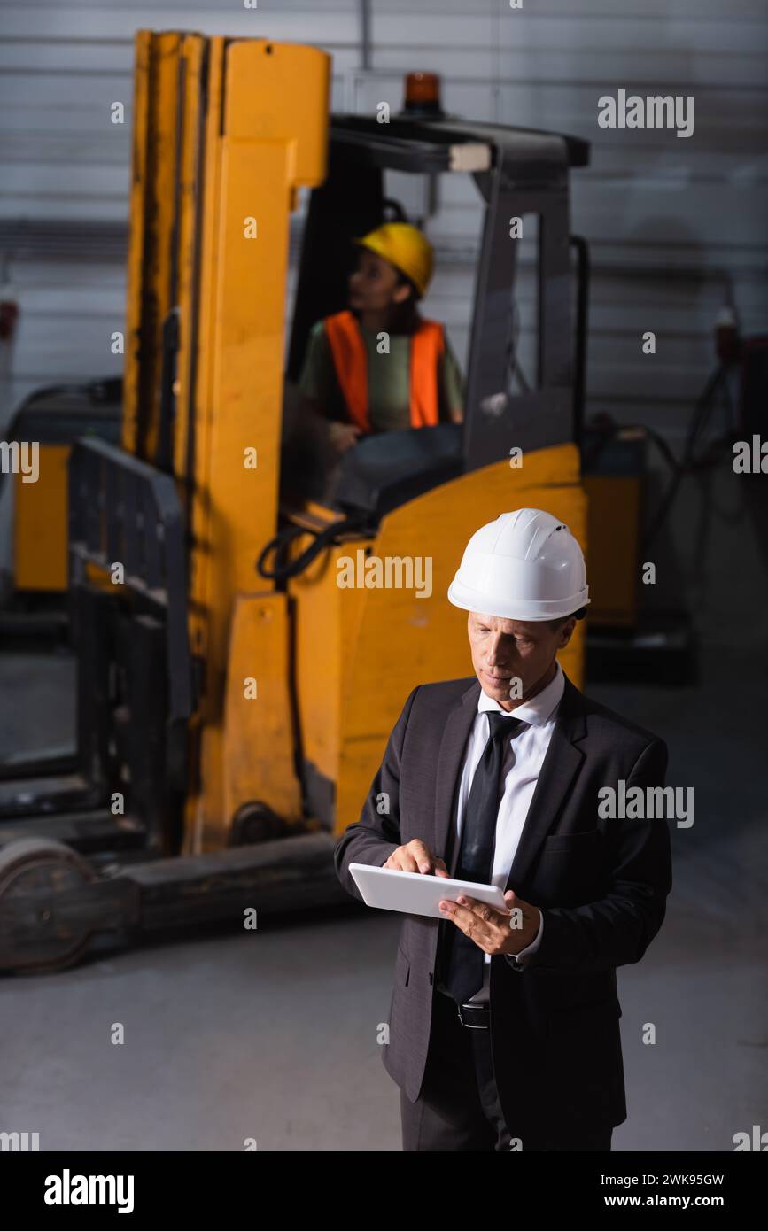 responsabile di magazzino di mezza età in abbigliamento aziendale e elmetto con tablet durante la pianificazione della logistica Foto Stock