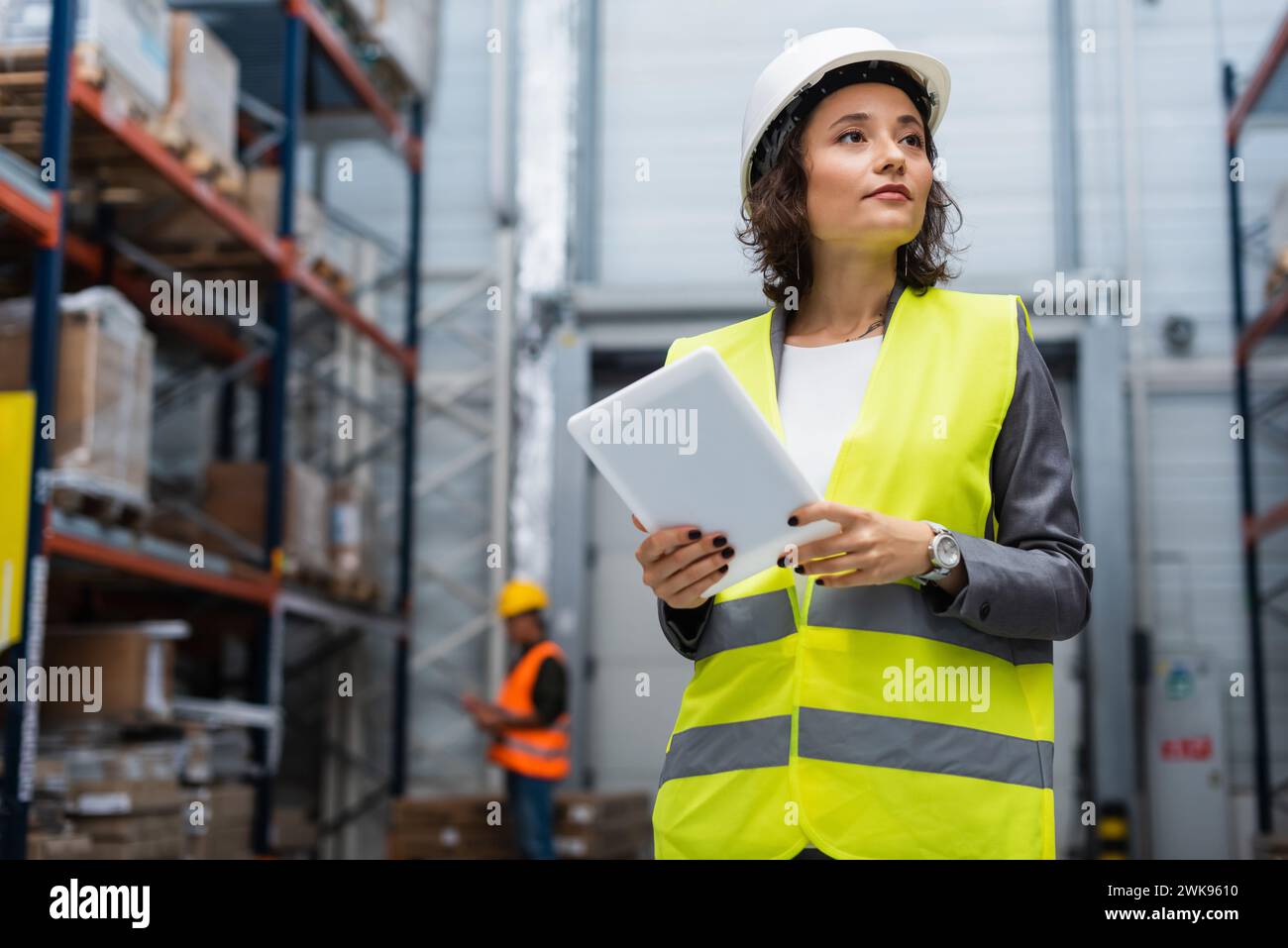 supervisore di magazzino donna in elmetto e giubbotto di sicurezza che tiene in mano un tablet digitale durante il lavoro Foto Stock