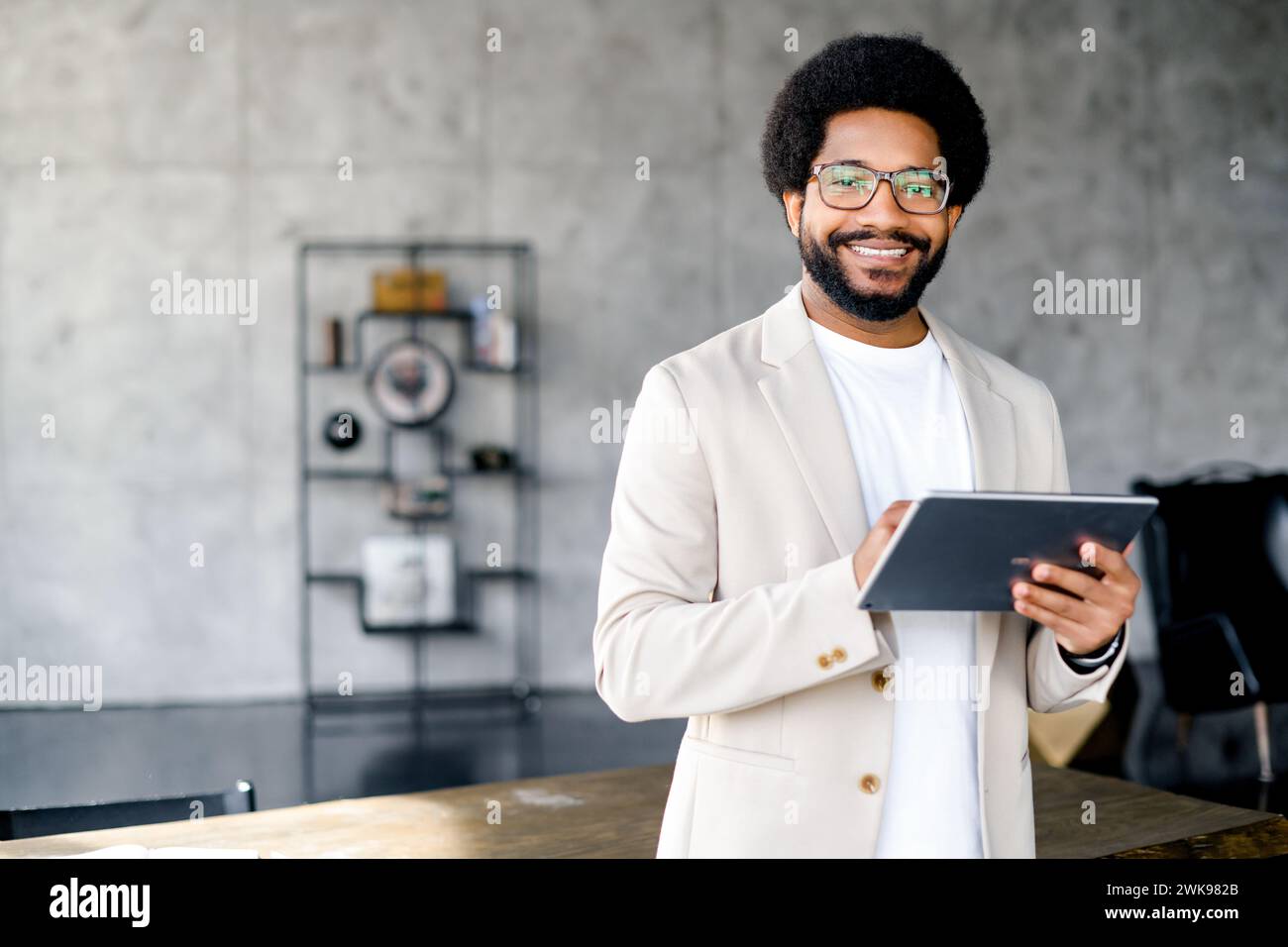 Giovane professionista in ufficio con un tablet, in piedi su uno sfondo industriale, un ambiente di lavoro moderno e creativo. Il suo sorriso suggerisce un approccio amichevole agli affari Foto Stock