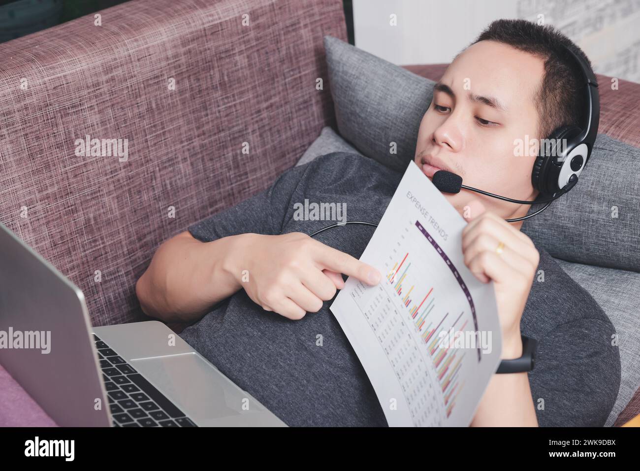 Giovane uomo sdraiato sul divano e videochiama i colleghi con un computer portatile in ufficio. Foto Stock