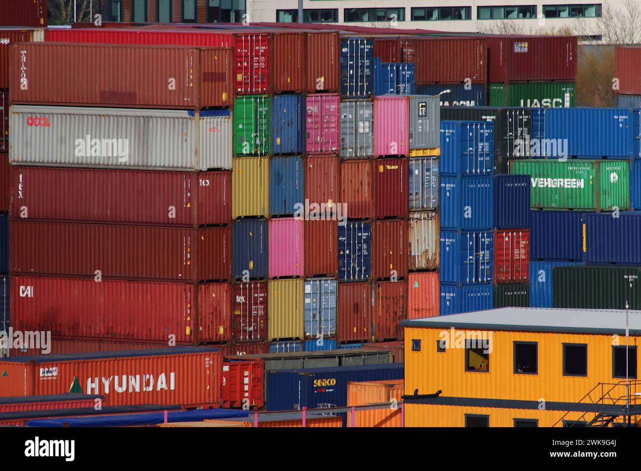 Città dei container - un'alta pila di container per spedizioni multiple sminuisce un edificio giallo di uffici a tre piani sulla banchina di Southampton, aprile 2023. Foto Stock