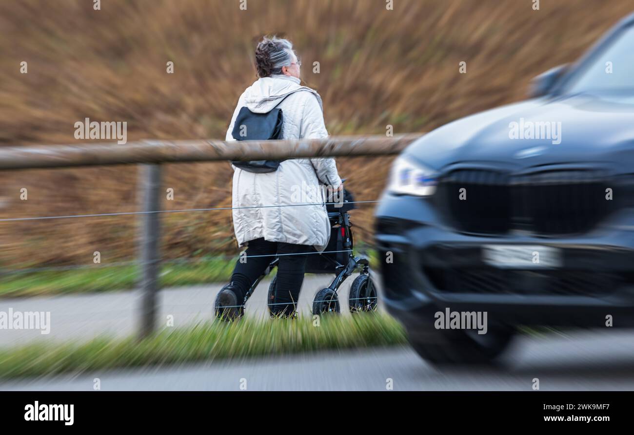 Eine Rentnerin läuft an einem Rollator der Strasse entlang. Davor kreuzt ein SUV der Marke BMW. (Oberglatt, Schweiz, 14.01.2023) Foto Stock