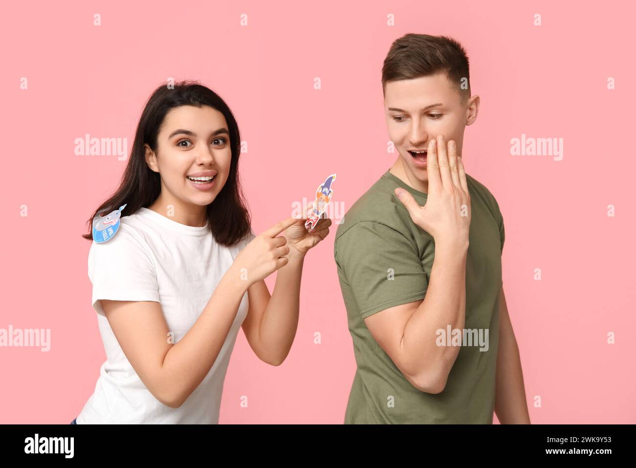 Giovane donna che attacca il clown di carta sulla schiena della sua amica su sfondo rosa. Festa del giorno dello scemo d'aprile Foto Stock