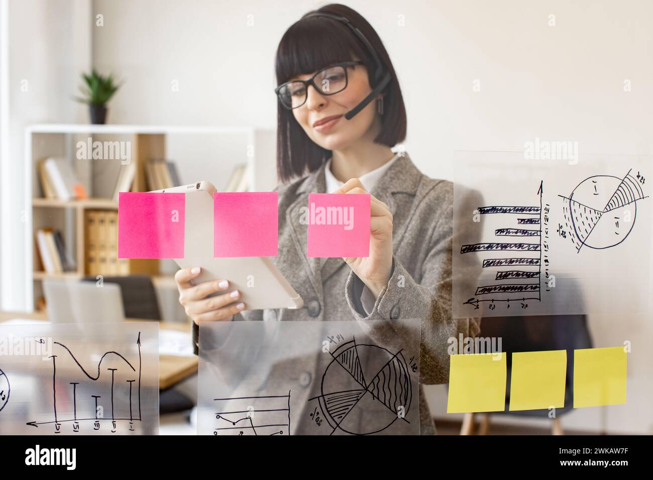 Donna caucasica che tiene il tablet in mano mentre si trova vicino a un tabellone da ufficio trasparente. Imprenditrice donna in cuffia che parla con il partner commerciale tramite Foto Stock