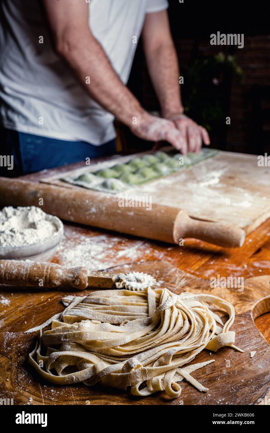 Primo piano di pasta fatta a mano chiamata fettuccine, lasciata riposare. Lo chef sullo sfondo, fuori dal fuoco, sta completando la preparazione dei ravioli verdi. Foto Stock