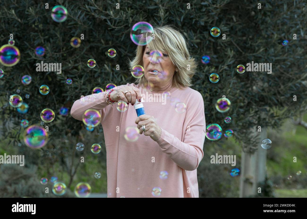 Una donna anziana con un maglione rosa crea gioiosamente bolle di sapone, catturando un momento di puro divertimento e giocosità durante una riunione femminile. Foto Stock