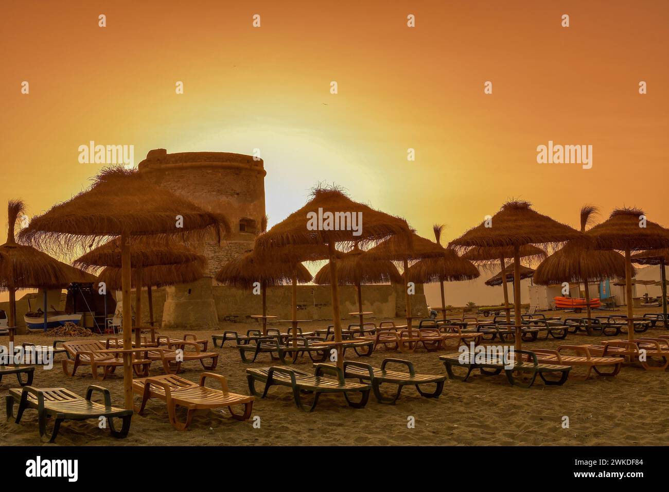 Tramonto dorato sulla tranquilla spiaggia di Cabo de Gata, con una storica torre di guardia e ombrelloni di paglia. Foto Stock